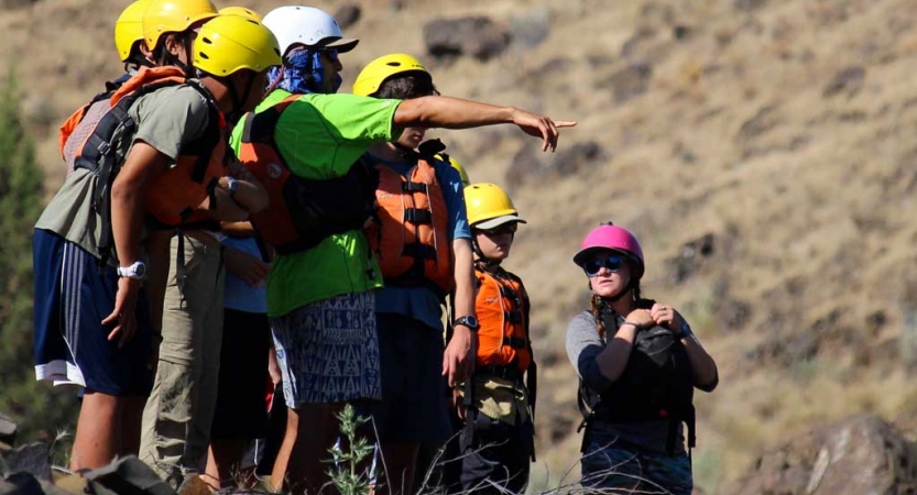 A group of people wearing helmets and life jackets stand on land. One of them points outward.